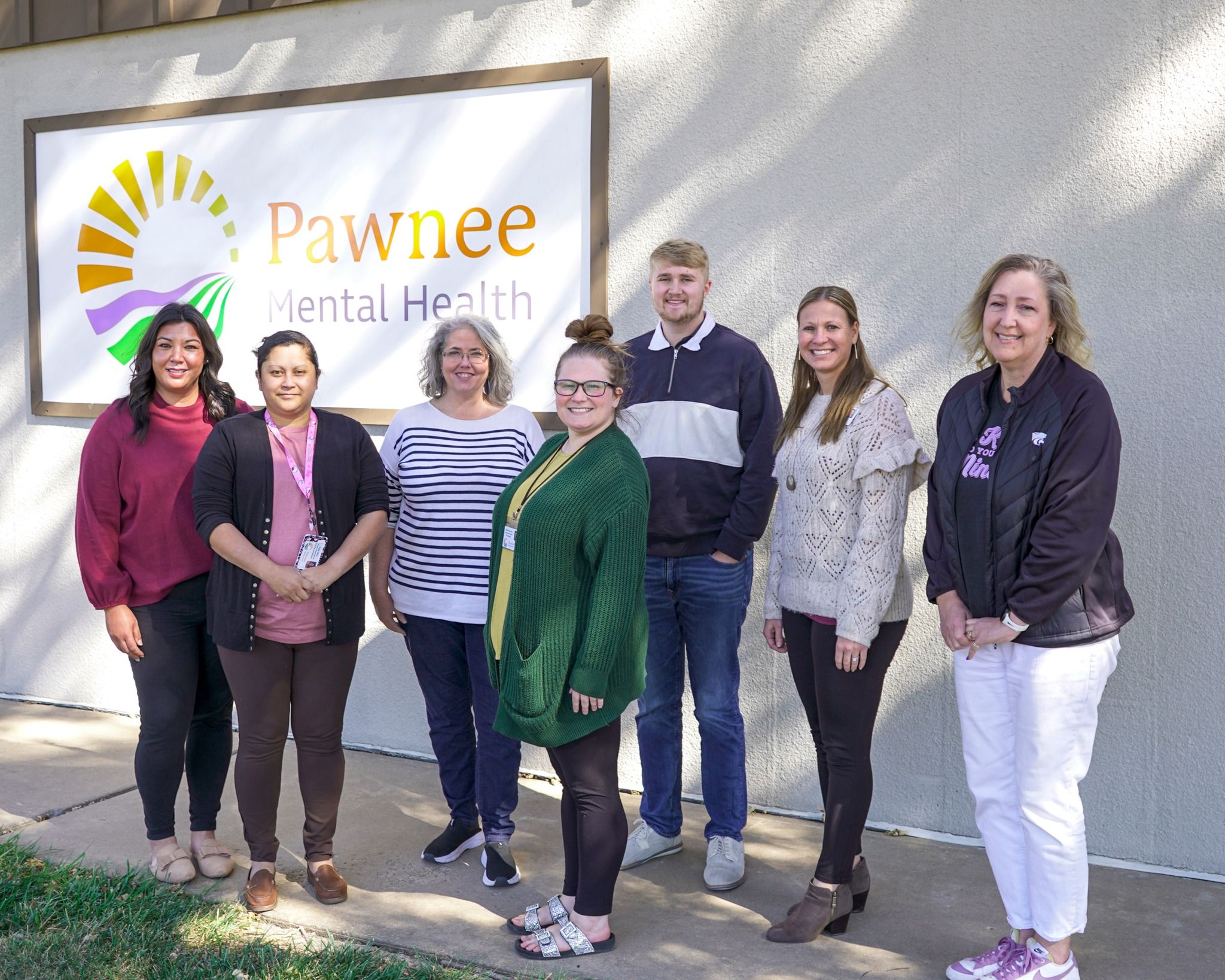 Seven diverse individuals stand smiling in front of "Pawnee Mental Health," exuding warmth, support, and community.