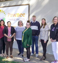 Seven diverse individuals stand smiling in front of "Pawnee Mental Health," exuding warmth, support, and community.
