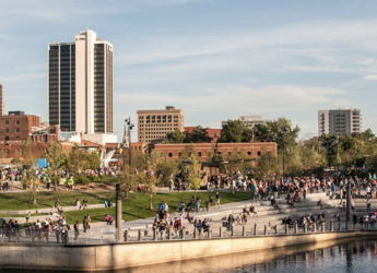 Vibrant urban park by a river, with people enjoying outdoor activities amidst green grass and a city skyline. Happy atmosphere.