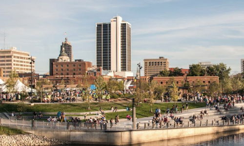Vibrant urban park by a river, with people enjoying outdoor activities amidst green grass and a city skyline. Happy atmosphere.