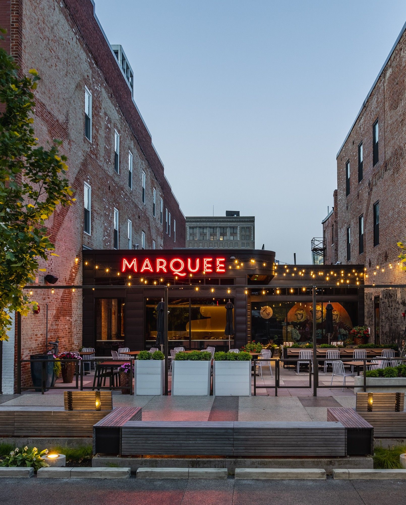 Lively outdoor venue "MARQUEE" with warm lighting, brick buildings, greenery, and inviting seating for social gatherings.