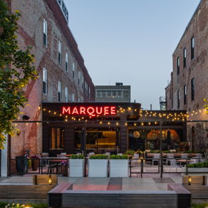 Lively outdoor venue "MARQUEE" with warm lighting, brick buildings, greenery, and inviting seating for social gatherings.
