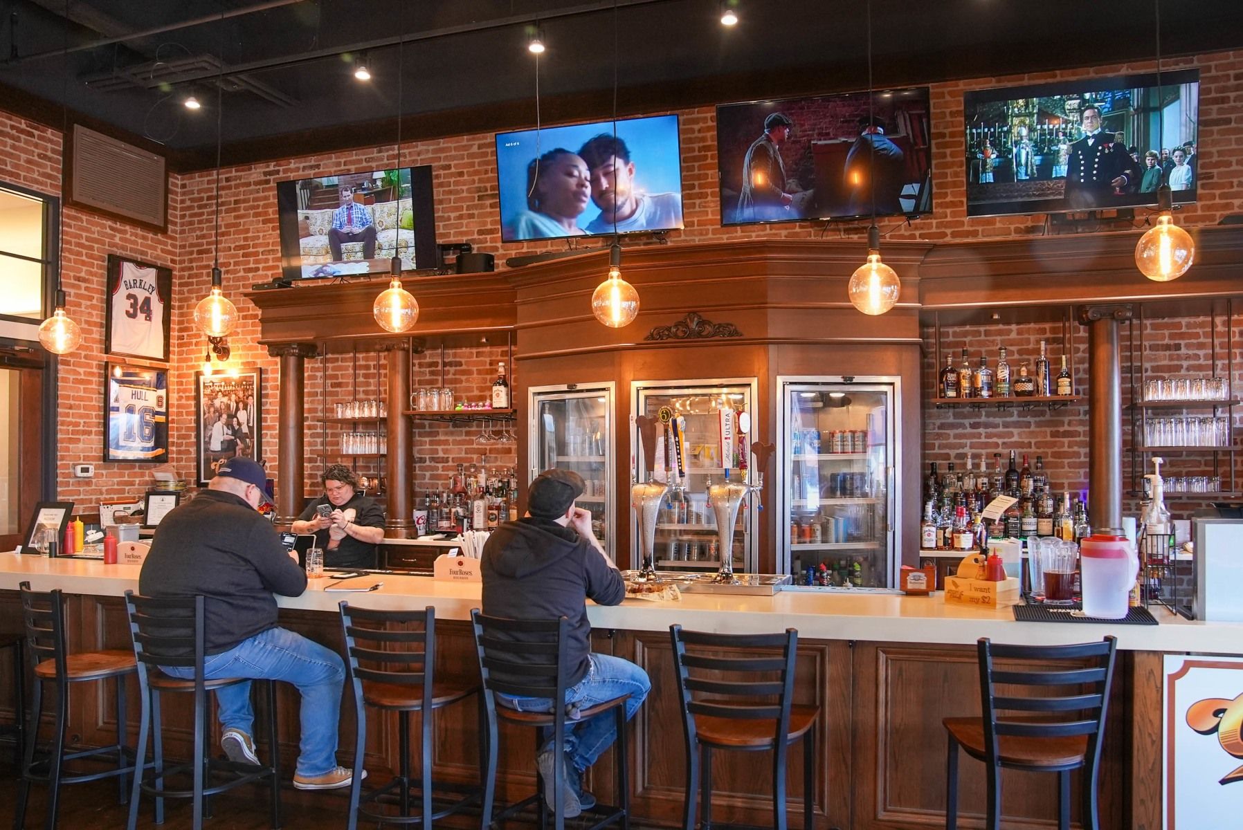 Cozy pub with exposed brick, warm lighting, patrons at the bar, and TVs, creating a vibrant, friendly atmosphere for socializing.