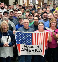 Diverse group in work attire holding "MADE IN AMERICA" sign, expressing pride, camaraderie, and joy in a manufacturing setting.