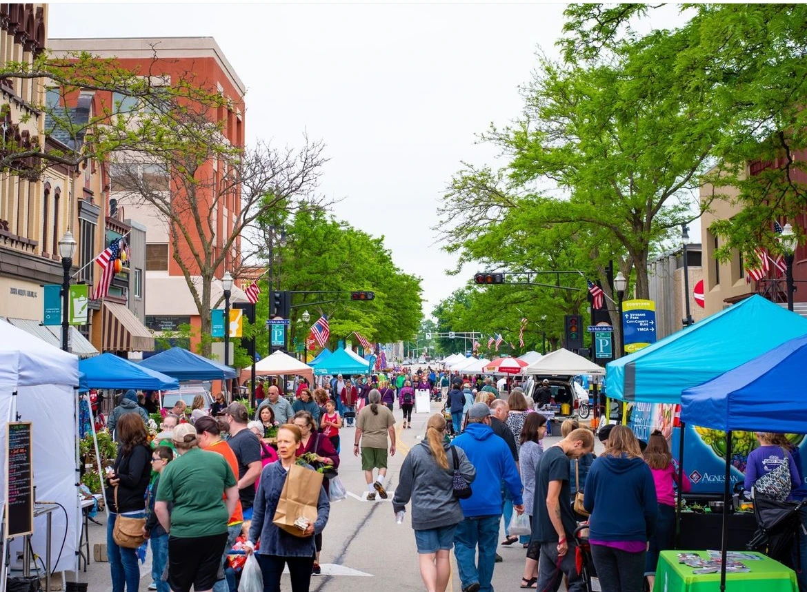 A lively street fair bustling with people, colorful booths, and trees, evoking a sense of community and excitement.