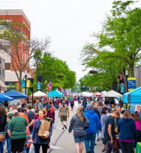 A lively street fair bustling with people, colorful booths, and trees, evoking a sense of community and excitement.