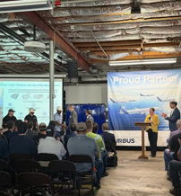Event gathering in an industrial setting with an Airbus banner; audience engaged, reflecting pride and community in aviation.