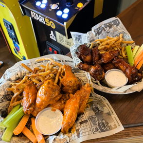 Two plates of saucy chicken wings with fries and celery, set on newsprint, beside a vintage arcade game, evoking fun times.