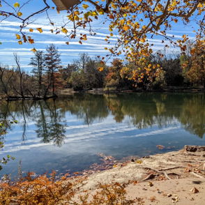 A tranquil autumn landscape with colorful trees reflecting in calm water under a partly cloudy sky.