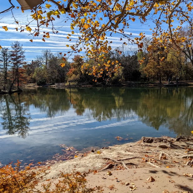 A tranquil autumn landscape with colorful trees reflecting in calm water under a partly cloudy sky.