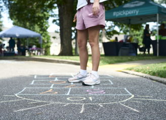 A young person plays hopscotch on pavement, evoking nostalgia, joy, and community spirit at a lively outdoor event.