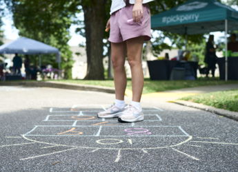 A young person plays hopscotch on pavement, evoking nostalgia, joy, and community spirit at a lively outdoor event.