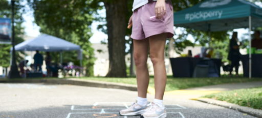 A young person plays hopscotch on pavement, evoking nostalgia, joy, and community spirit at a lively outdoor event.