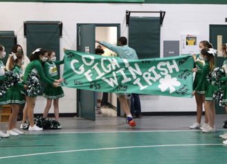 Cheerleaders in green uniforms hold a "GO FIGHT WIN IRISH" banner in a festive gym, radiating school spirit and excitement.