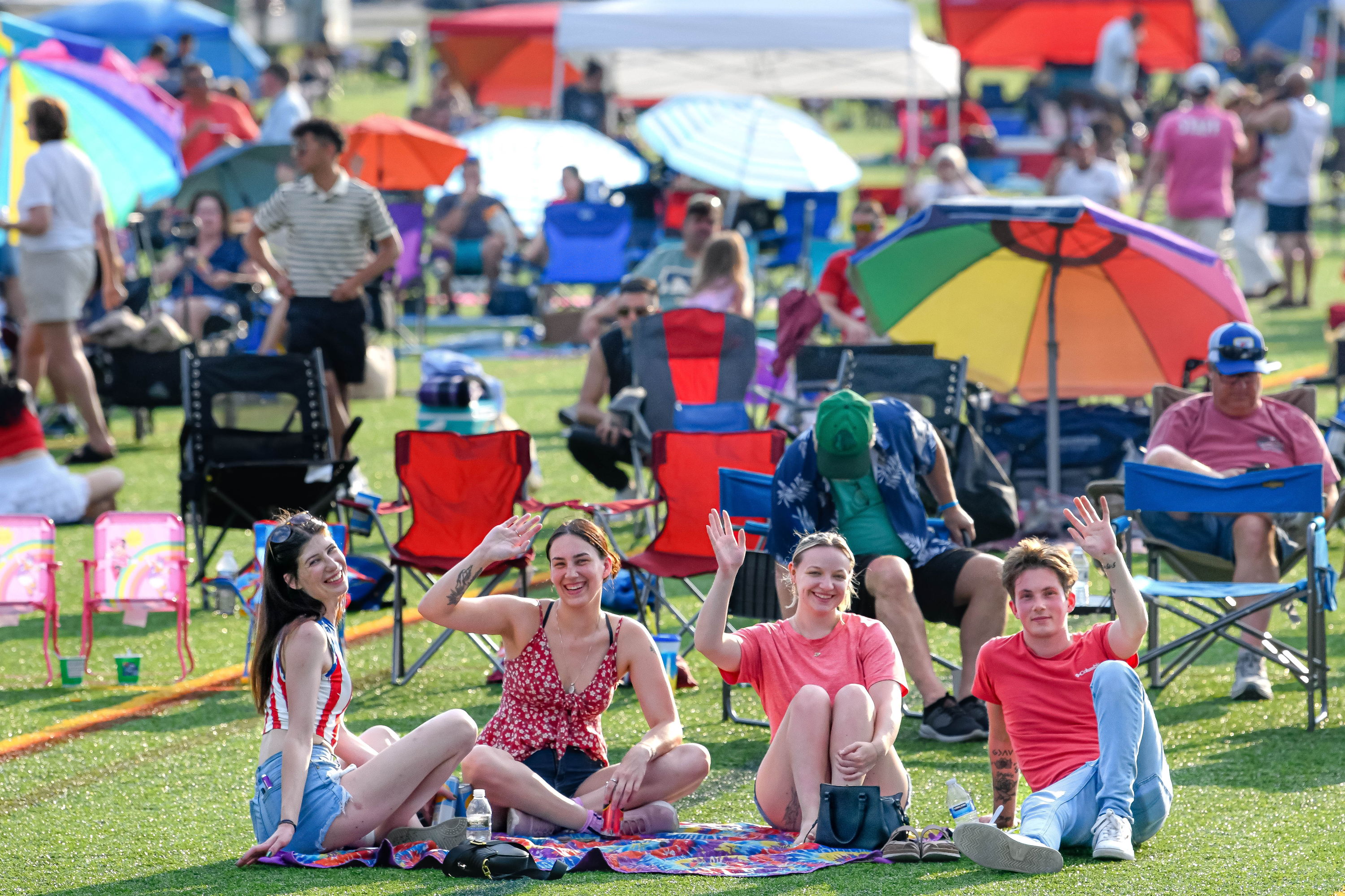 Four young women sit on a colorful blanket at a lively outdoor festival, smiling and enjoying a sunny, joyful atmosphere.