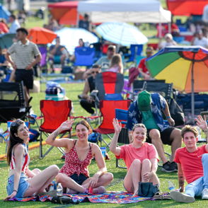 Four young women sit on a colorful blanket at a lively outdoor festival, smiling and enjoying a sunny, joyful atmosphere.