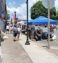 Lively street scene at a festival with vendors, people strolling, a bright sky, and a joyful community atmosphere.