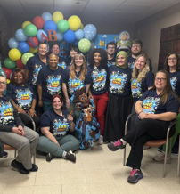 Group of individuals in colorful t-shirts celebrate joyfully under a balloon arch, embodying community and togetherness.