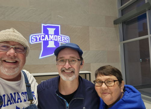 Three smiling individuals in a bright space with a "SYCAMORES" sign, radiating joy and camaraderie at a school event.