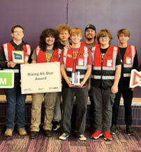 Young people in red vests celebrate an achievement, holding awards and signs, showcasing teamwork and joy at a ceremony.