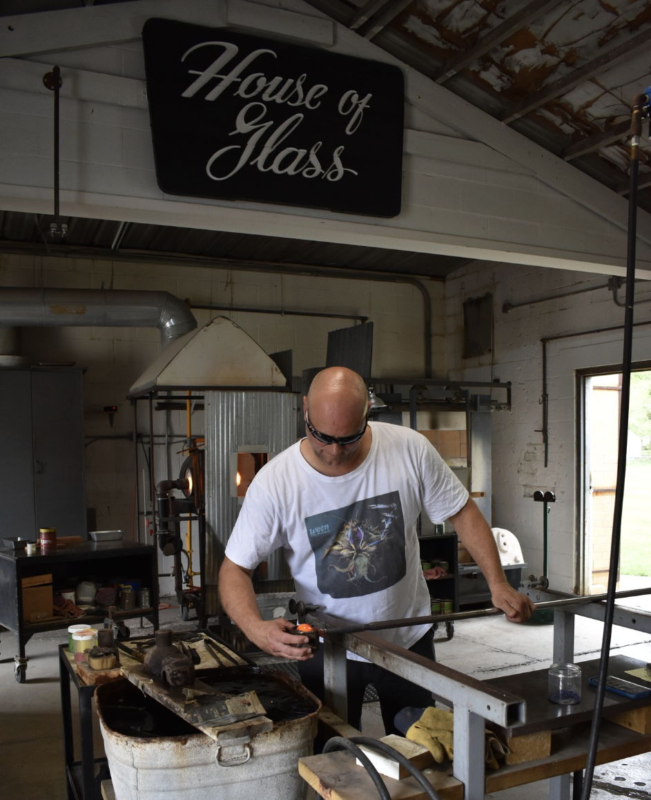 A glass artist works intently in the "House of Glass," showcasing dedication and creativity in a cozy, well-lit workshop.
