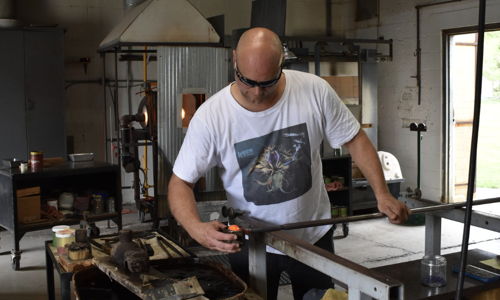 A glass artist works intently in the "House of Glass," showcasing dedication and creativity in a cozy, well-lit workshop.