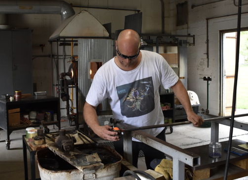 A glass artist works intently in the "House of Glass," showcasing dedication and creativity in a cozy, well-lit workshop.