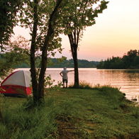 Lakeside camping at sunset, with a tent, trees, calm water, and a person in contemplation, evokes peace and connection to nature.