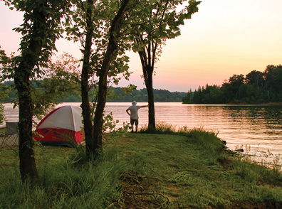Lakeside camping at sunset, with a tent, trees, calm water, and a person in contemplation, evokes peace and connection to nature.