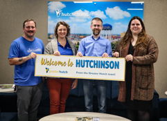 Four smiling people hold a "Welcome to HUTCHINSON" sign, creating a warm, inviting atmosphere in a community setting.