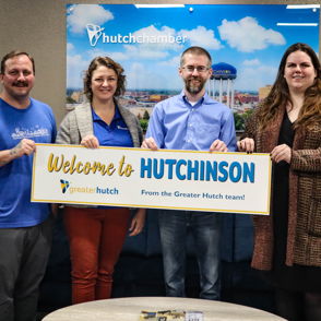 Four smiling individuals hold a sign reading "Welcome to HUTCHINSON" in a cheerful, community-focused environment.
