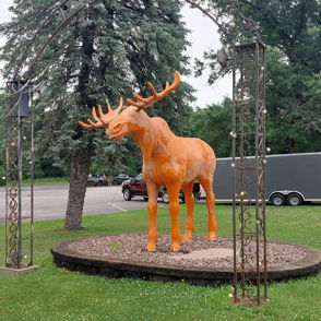 An orange moose sculpture with antlers stands on a stone platform under a decorative lit arch, surrounded by grass and trees.