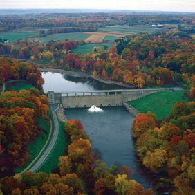 Scenic autumn landscape with a river, colorful trees, a dam, and an overcast sky, evoking tranquility and natural beauty.