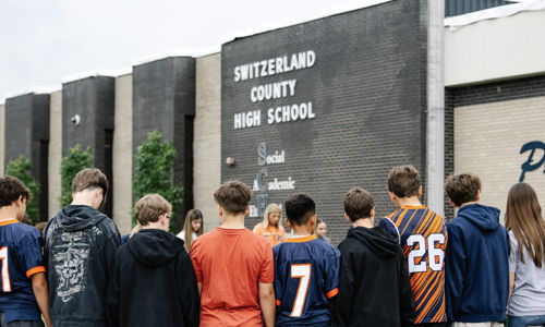 Students in jerseys stand united in front of a high school, exuding camaraderie and anticipation for a meaningful event.