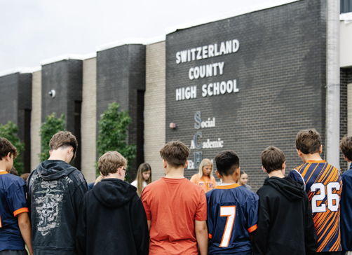 Students in jerseys stand united in front of a high school, exuding camaraderie and anticipation for a meaningful event.