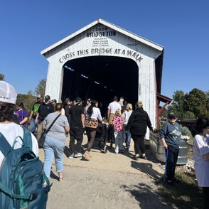 A lively crowd enters a rustic covered bridge under a clear sky, evoking a sense of community and adventure.
