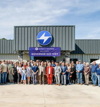 Group photo outside an industrial building with trucks; diverse people, formal attire, celebrating community and leadership event.