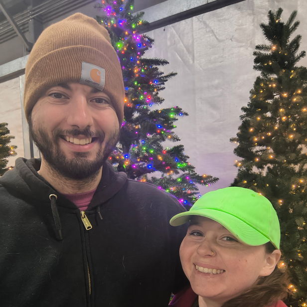 Couple smiling by festive Christmas trees, radiating joy and warmth in cozy jackets, celebrating the holiday spirit together.