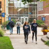 Campus scene with students walking, chatting in a vibrant, green setting, showcasing community and a lively atmosphere.