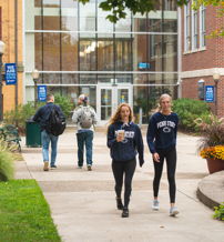 Campus scene with students walking, chatting in a vibrant, green setting, showcasing community and a lively atmosphere.