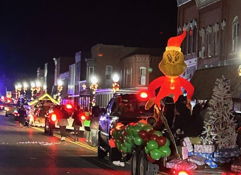 Festive nighttime parade scene with decorated vehicles, balloons, and a Grinch inflatable, evoking joy and community spirit.