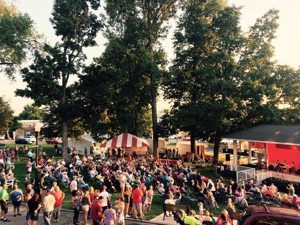 A vibrant outdoor festival with a large crowd, trees, a red stage, and a joyful atmosphere of celebration and community.