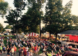 A vibrant outdoor festival with a large crowd, trees, a red stage, and a joyful atmosphere of celebration and community.