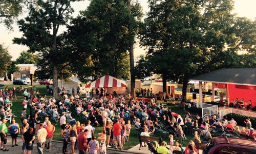 A vibrant outdoor festival with a large crowd, trees, a red stage, and a joyful atmosphere of celebration and community.