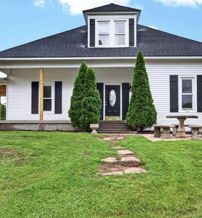 White house with black roof on a green lawn, pathway of stepping stones, shrubs, and outdoor seating evokes warmth and tranquility.