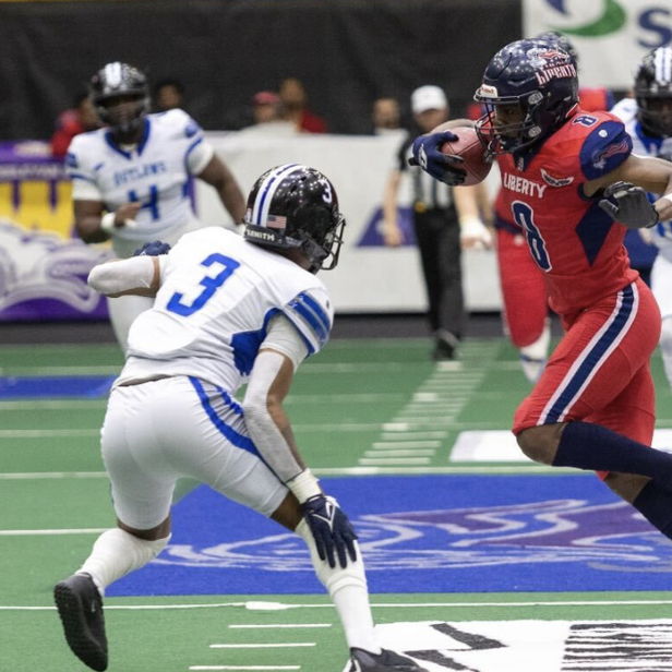 Two football players, one in red and the other in white/blue, clash in an exciting indoor game moment, showcasing intensity.
