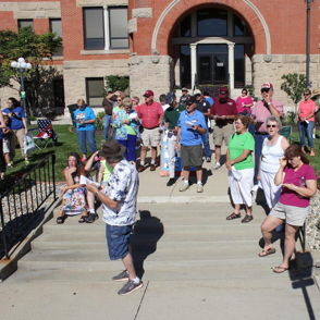 A diverse crowd in bright summer clothing gathers outside a building for a festive event, radiating excitement and camaraderie.