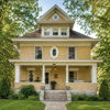 Charming yellow house with large windows, gable roof, surrounded by lush greenery, evokes comfort and nostalgia.