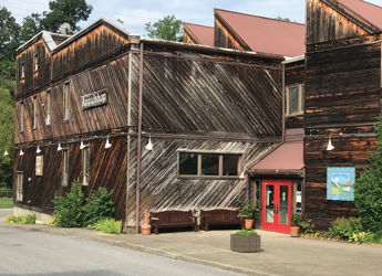 Rustic wooden building with a warm, inviting vibe, featuring a red door, green shrubs, and a charming, weathered design.