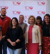 A diverse group celebrates in front of a "100" backdrop, expressing joy and camaraderie, marking a significant achievement.